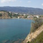 A coastal town by a calm blue sea with hills in the background.