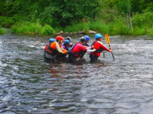 Four people wearing helmets and life jackets wade through a shallow river.