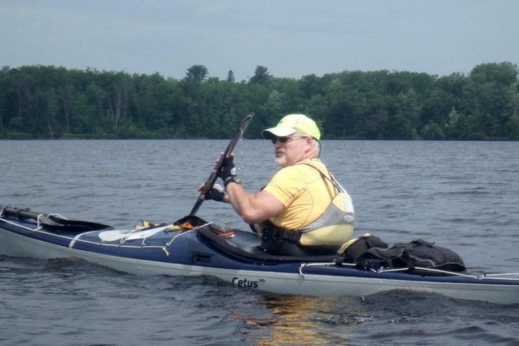 27929989271_72c4da83a7_o-2-1024x683 A person kayaking on a calm lake wearing a yellow life jacket and cap.