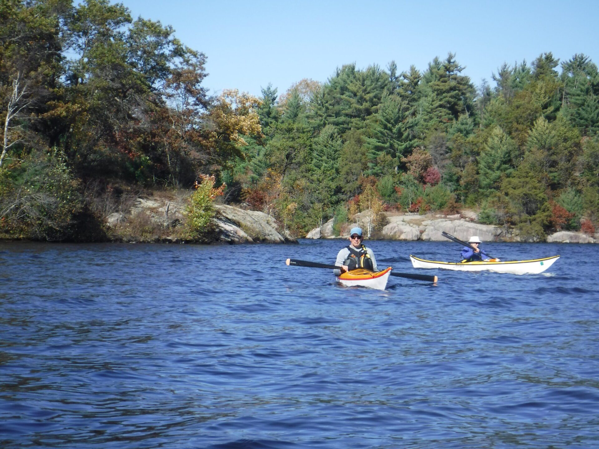 DSCF0960-scaled (1) Two people kayaking on a river surrounded by trees.