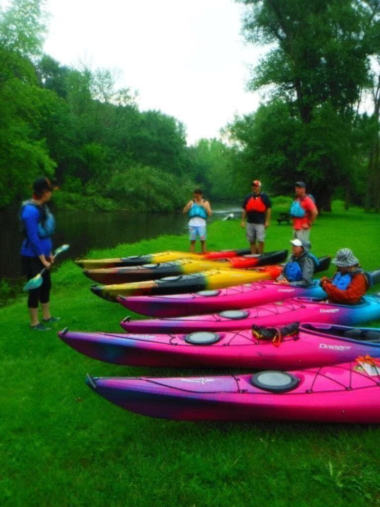 DSCF1838-768x1024 (1) A group of people with colorful kayaks on green grass.