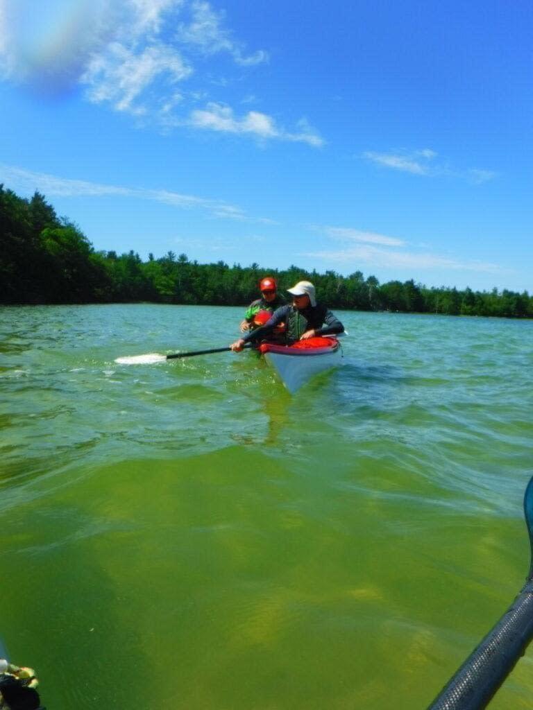 DSCF1891-768x1024 Two people kayaking on a calm lake under a blue sky.