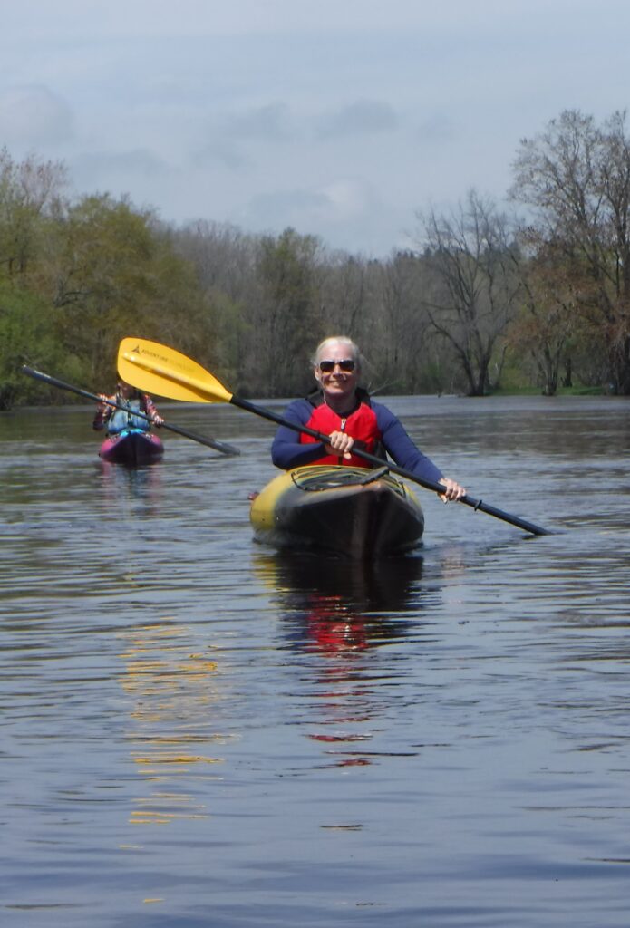 DSCF4125-695x1024 Two people kayaking on a calm river surrounded by trees.