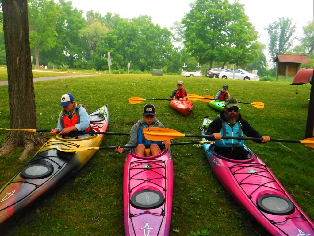 DSCF4218-1024x768 Five people sitting in colorful kayaks on grass near trees.