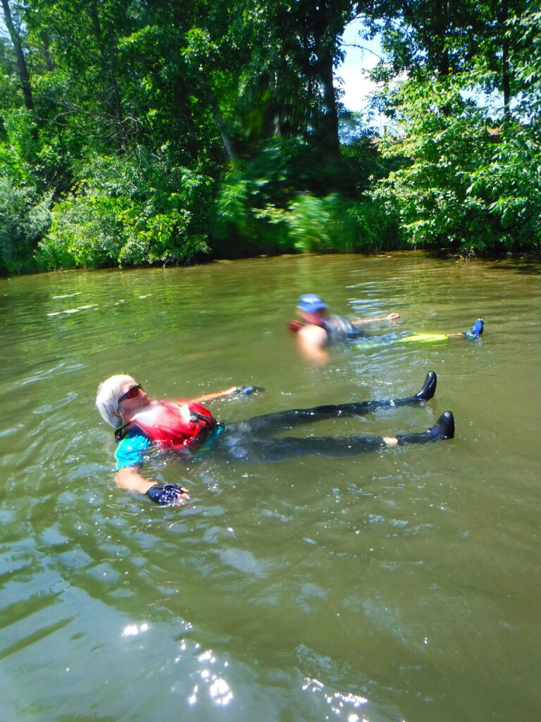 DSCF4518-768x1024 Two people floating on their backs in a river surrounded by greenery.