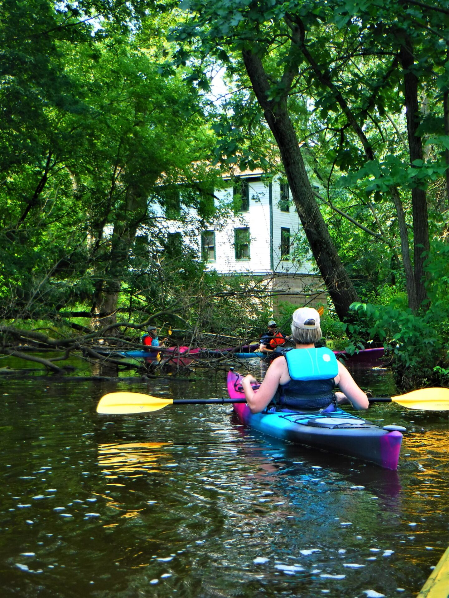 Two people kayaking on a river near a white house in a forest.