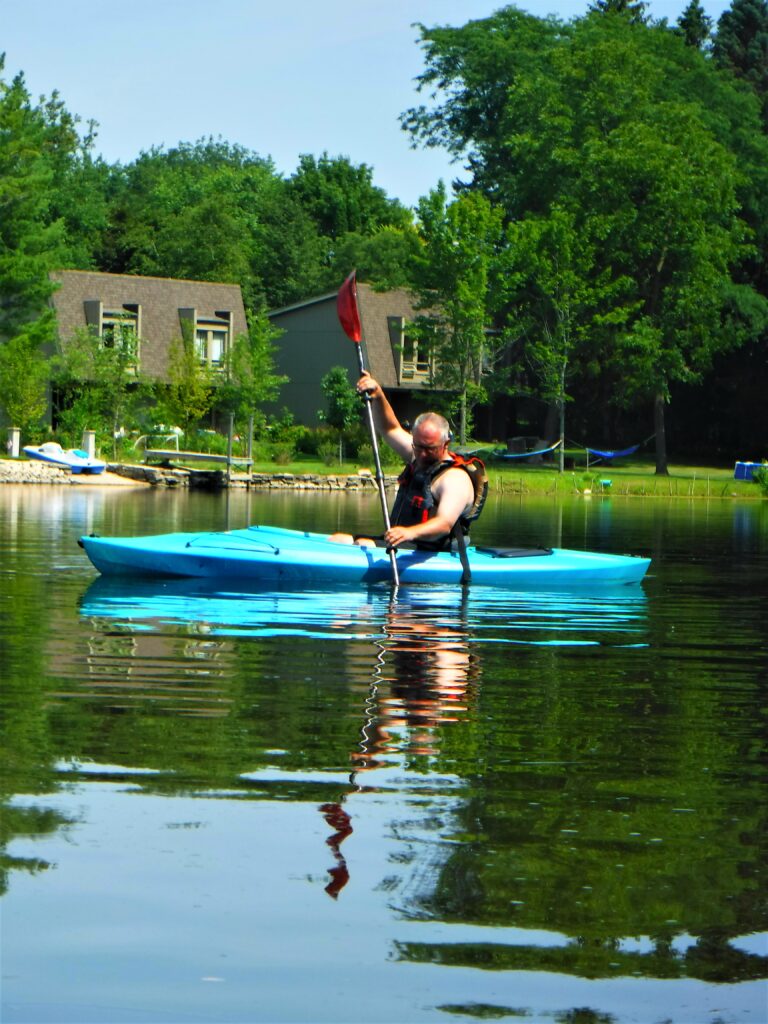 DSCF4673-768x1024 A person kayaking on a calm lake near a cabin.