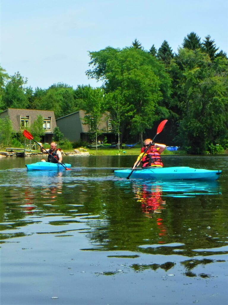 DSCF4675-768x1024 Two people kayaking on a calm lake near green trees.