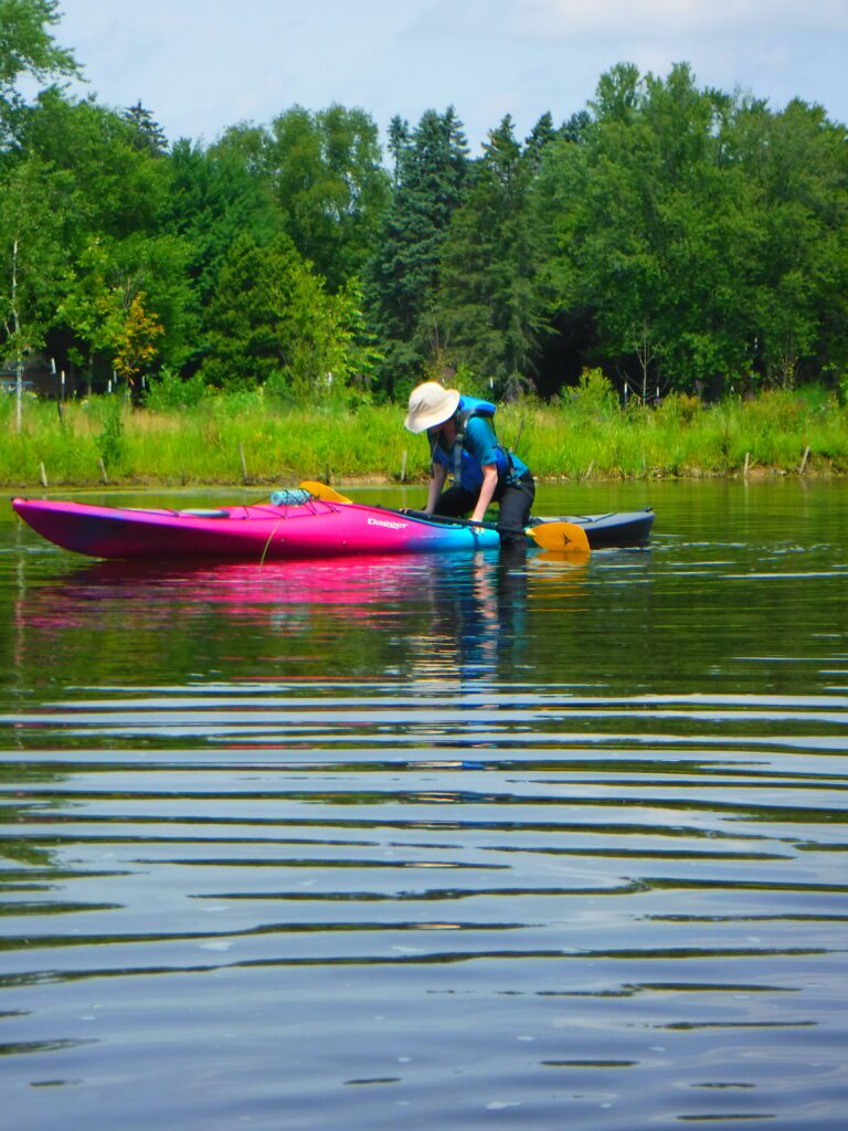 DSCF4679-768x1024 A person in a blue jacket and hat paddling a pink kayak on calm water.