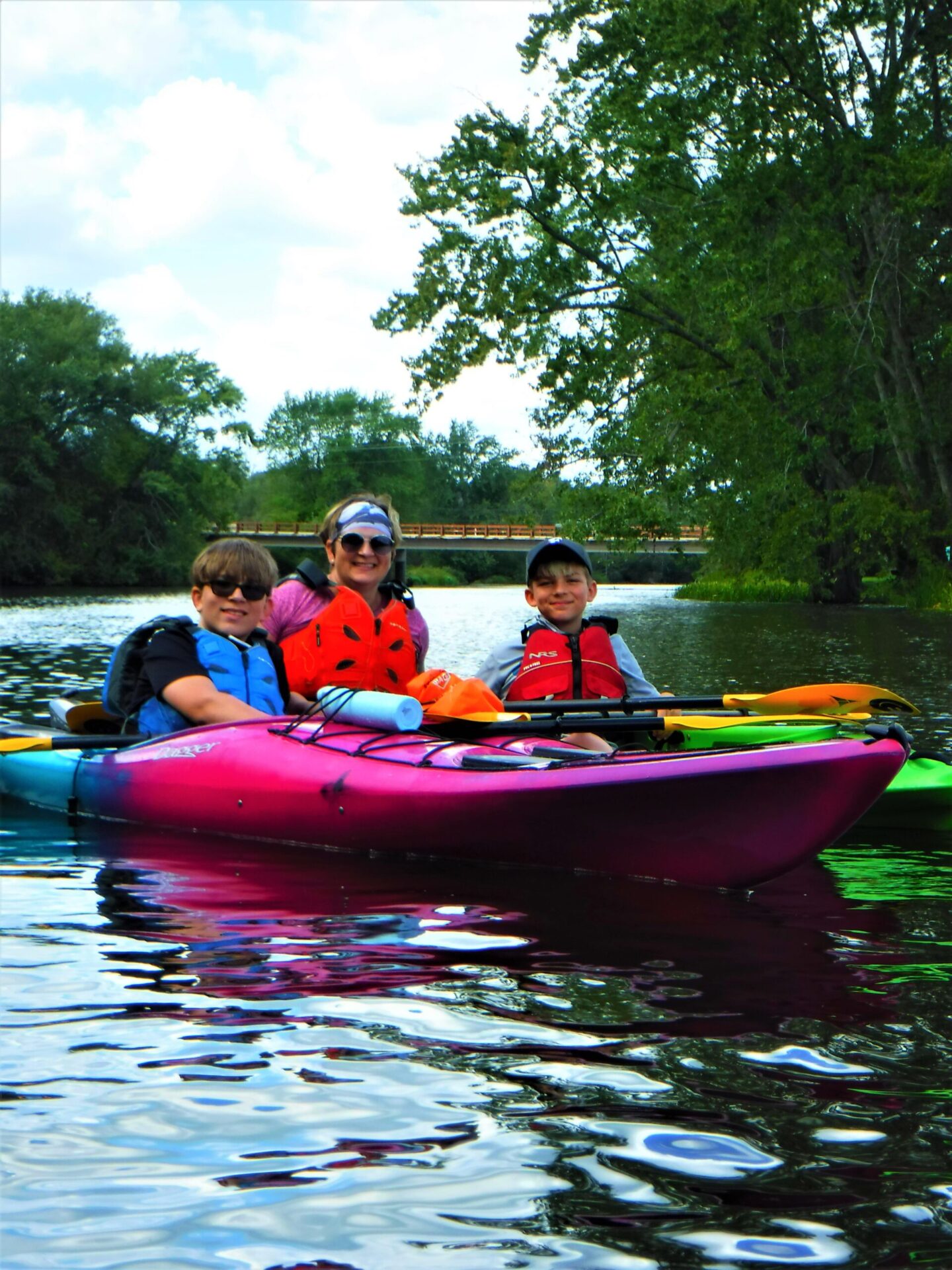 DSCF4926-scaled Three people kayaking on a calm river surrounded by green trees.