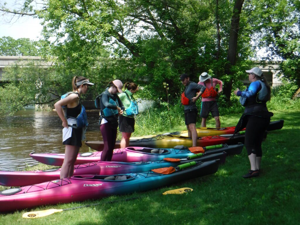 DSCF7418-1024x768 A group of people standing near colorful kayaks on grass.