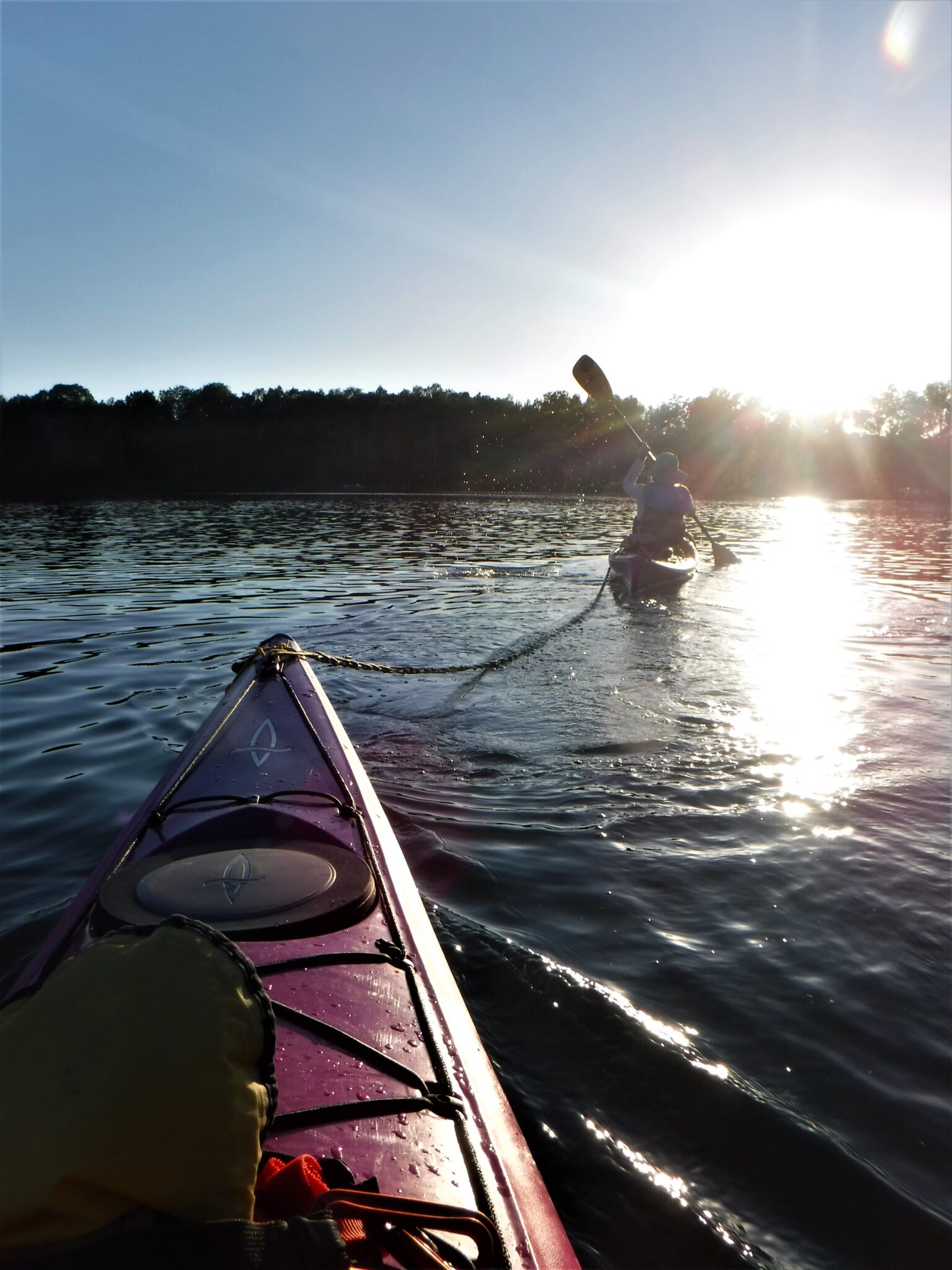 DSCF7446-1536x2048 A kayak on calm water with a person fishing in the background at sunset.
