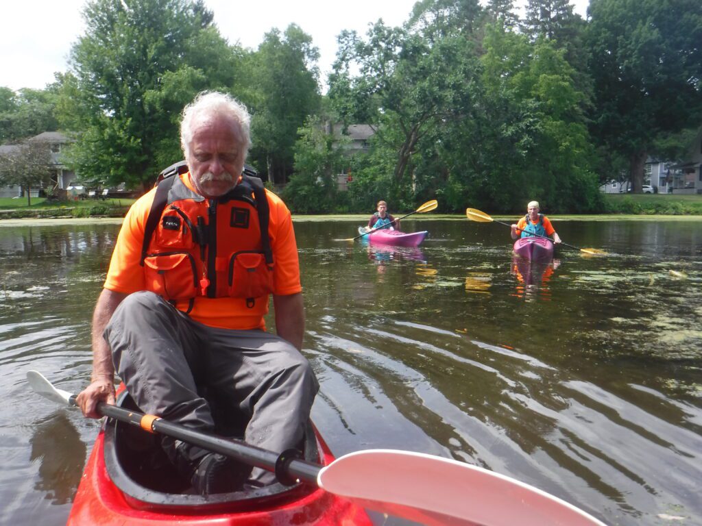 DSCF7929-1024x768 A man in an orange life jacket paddling a kayak with two other kayakers in the background.